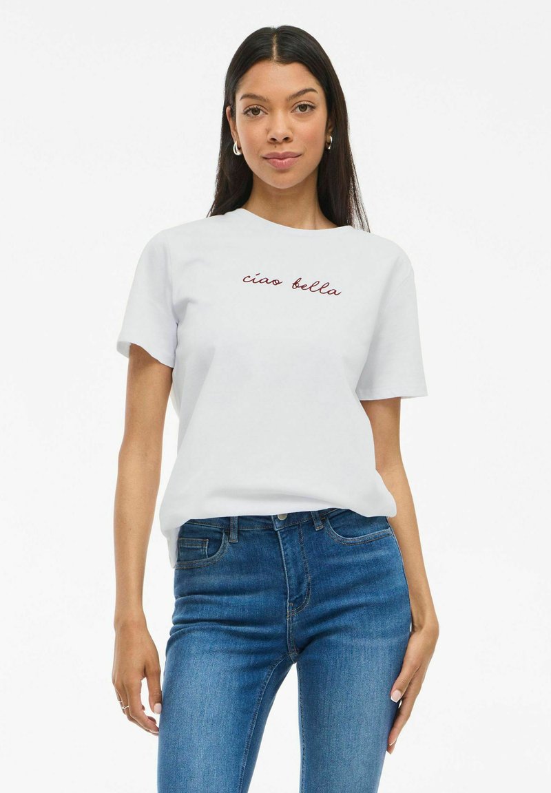 T-shirt en coton blanc à manches courtes, avec un texte en cursive rouge "ciao bella" sur la poitrine. Assorti avec un jean en denim bleu.