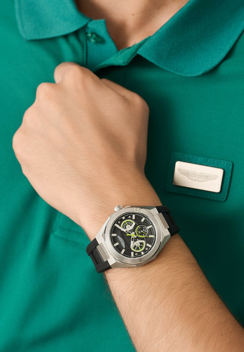 Orologio sportivo argento con cinturino in gomma nera, quadrante nero e dettagli verdi. Indossato su una mano accanto a una maglietta polo verde e un'etichetta.