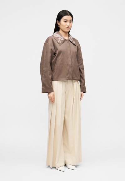 Γυναίκα με καφέ μπουφάν με κεντημένο γιακά, εκρού φαρδιά πλισέ παντελόνια, λευκά μυτερά παπούτσια, στέκεται μπροστά σε ένα απλό φωτεινό φόντο.