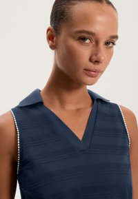 Mornarsko modra brezrokavnik s prihajajočim izrezom v obliki črke V, teksturiranimi črtami in ovratnikom, ki vključuje majhne biserne okraske ob ramah.