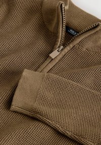 Hnedý sveter so zapínaním na zips s textúrou v štýle vafľového pletenia. Má vysoký golier a hladký zips s matným povrchom.