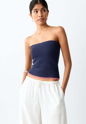 Jeune femme portant un haut sans bretelles en crochet bleu marine avec une bordure rose et un pantalon blanc à cordon, debout les mains dans les poches, regardant vers la droite.