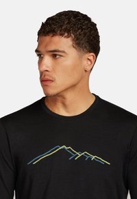 Jonge man met kort krullend haar, gekleed in een zwarte shirt met een blauw en geel berglijnontwerp, kijkend naar links tegen een witte achtergrond.
