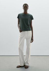 Donkere groene T-shirt met korte mouwen, relaxed fit, gecombineerd met witte wijde jeans. Model draagt bruine sandalen en staat voor een lichte achtergrond.