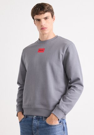Gray sweatshirt with a crew neck, ribbed cuffs, and a red rectangular patch labeled "HUGO" on the chest. Worn with blue jeans.