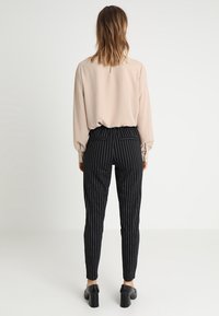Beige Bluse mit langen Ärmeln, kombiniert mit schwarzen gestreiften Hosen. Die Hose hat einen Bund und zwei Gesäßtaschen. Schwarze High Heels.