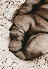 Rjavo teksturirano flisasto obleko z zadrgo spredaj, mehke manšete in pokrivala za noge, ki počivajo na lahki pleteni odeji z vzorcem listov.