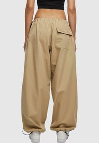 Pantalons beige à jambes larges avec une taille élastique, poche arrière et poignets ajustables, fabriqués en tissu léger. Portés avec des baskets blanches.