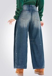 Jeans in denim blu sbiadito a gamba larga con vita alta, dotati di due tasche posteriori e cuciture a contrasto. Abbinati a calzature marroni.