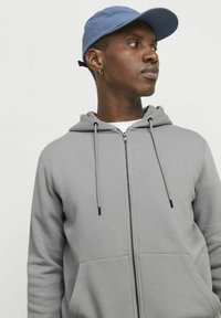 Graue Zip-Hoodie mit einer Fronttasche, verstellbaren Kordeln und einer glatten Textur. Trägt eine hellblaue Kappe.