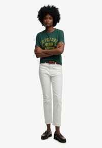 Zielona bawełniana koszulka z żółtym logo Superdry, białe dżinsowe spodnie i ciemnobrązowe mokasyny. Kręcone włosy, skrzyżowane ramiona, pozycja stojąca.