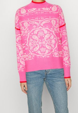 Lyserød sweater med hvid paisley-mønster, røde accenter på manchetter og krave, båret med lyseblå jeans.