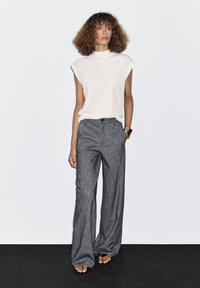 White, sleeveless top with a high neckline; paired with gray, wide-leg trousers featuring a subtle texture and front pockets. Flat sandals.
