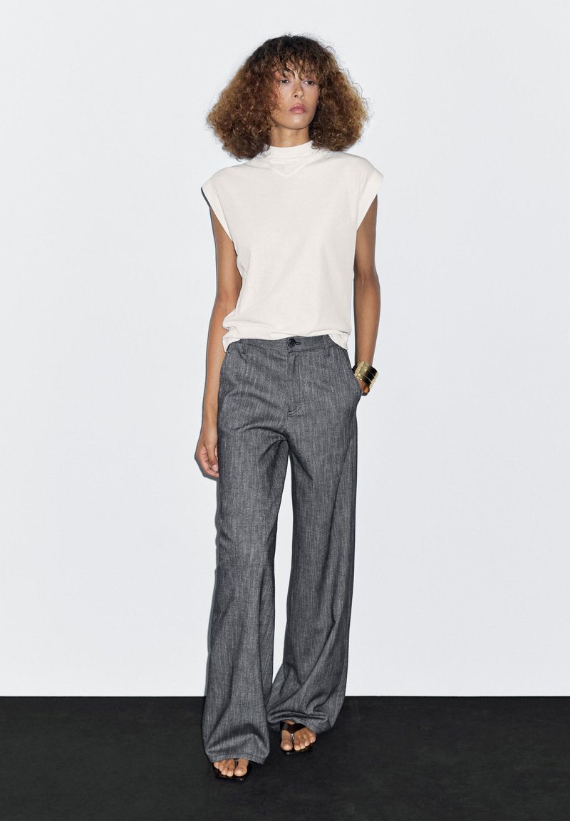 White, sleeveless top with a high neckline; paired with gray, wide-leg trousers featuring a subtle texture and front pockets. Flat sandals.