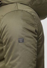 Olivgrön vadderad jacka med synliga sömmar. Har en fyrkantig metalllogg som är sydd på vänster arm. Slät textur.