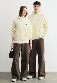 Δύο άτομα φορούν κρεμ φούτερ με κουκούλα και φαρδιά καφέ παντελόνια, στέκονται σε γκρι δάπεδο μπροστά από έναν λευκό τοίχο· το ένα κρατά μια μικρή μαύρη τσάντα.