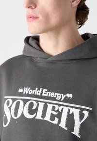 Šedá bavlnená mikina s kapucňou s predným dizajnom, na ktorom je text "World Energy Society" v tučných bielych písmenách a vykrojená kapucňa.