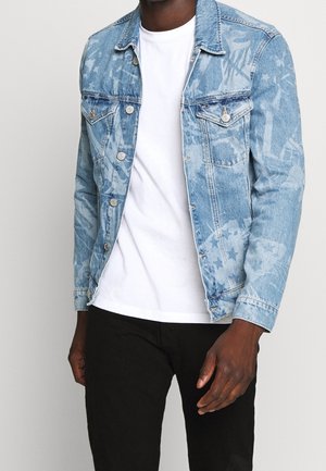 Man draagt een lichtblauwe spijkerjas met patroon over een wit T-shirt en zwarte broek, staand tegen een effen achtergrond.