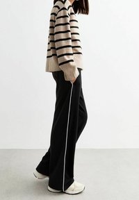 Pantalons larges noirs avec des rayures blanches sur les côtés, associés à un pull rayé beige et noir. La tenue comprend des baskets de couleur claire.