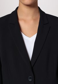 Blazer noir avec une fermeture à un bouton, des revers crantés et une texture de tissu lisse. Porté sur un haut blanc décolleté en V.