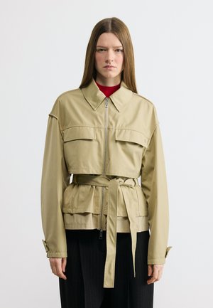 Γυναίκα που φοράει μπεζ σακάκι με ζώνη, μπροστινό φερμουάρ και τσέπες με καπάκια, συνδυασμένο με μαύρο παντελόνι και κόκκινο τοπ, στέκεται μπροστά σε απλό φόντο.