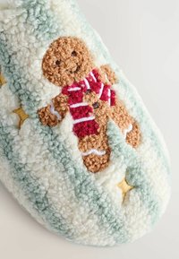 Papuci tricotati cu un om de turtă dulce care poartă o eșarfă roșie, pe un fundal moale în nuanțe de verde și crem, cu accente galbene.