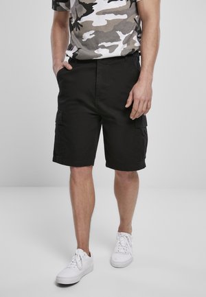 Uomo con pantaloncini cargo neri, scarpe da ginnastica bianche e t-shirt grigia con motivo mimetico, in piedi con le mani nelle tasche.