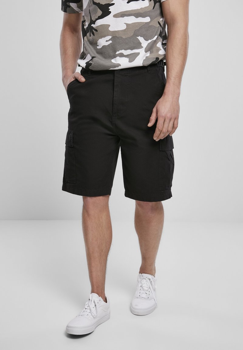 Homme portant un short cargo noir, des baskets blanches et un t-shirt gris à motif camouflage, debout les mains dans les poches.