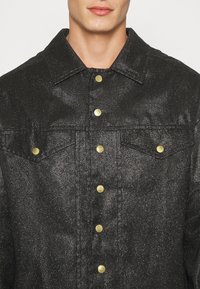 Svart denimjacka med en texturerad yta, guldknappar, två bröstfickor och en klassisk kragsdesign.