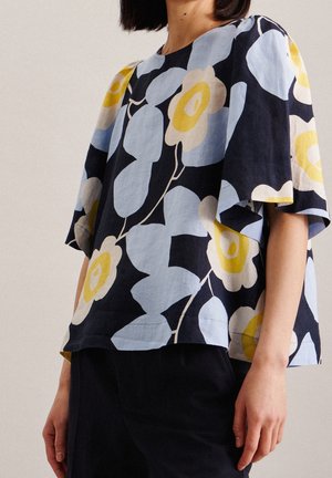 Kvinde iført en løs bluse med korte ærmer med stort blått og gult blomsterprint og sorte bukser, mod en ensfarvet lys baggrund.