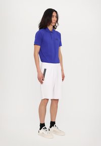 Blaues Polohemd mit kurzen Ärmeln und schwarzem Logo, kombiniert mit weißen Shorts mit schwarzen Streifen an der Seite und schwarzen Sportschuhen.