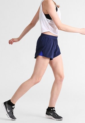 Femme marchant en short de sport bleu marine, brassière de sport noire, haut sans manches blanc et chaussures de course noires Nike sur un fond uni.
