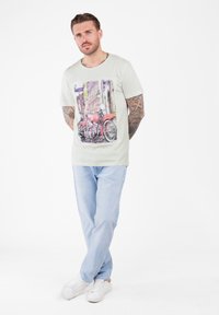 Hombre con los brazos tatuados que lleva una camiseta gráfica de motocicleta, jeans de mezclilla claros y zapatillas blancas, de pie con las piernas cruzadas.