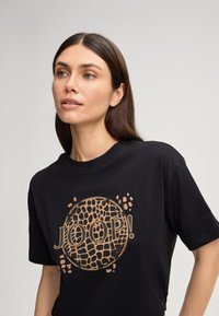 T-shirt en coton noir arborant un grand logo circulaire doré avec un motif texturé et le nom de la marque "JOOP!" bien en évidence.