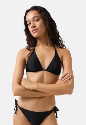 Junge Frau mit langen lockigen Haaren, die einen schwarzen Neckholder-Bikini trägt, mit verschränkten Armen vor einem schlichten hellgrauen Hintergrund stehend.