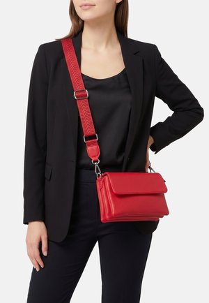 Donna che indossa un blazer nero e pantaloni, porta una borsa a tracolla in pelle rossa con una tracolla larga e regolabile.