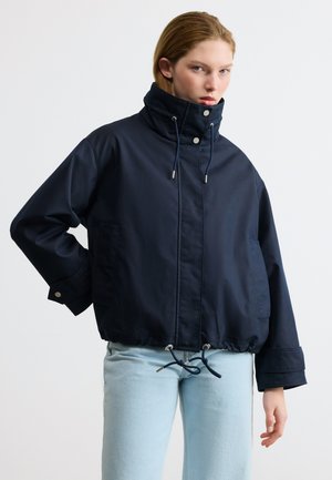 Jong persoon die een marineblauwe jas met hoge kraag en trekkoorden draagt, gecombineerd met lichtblauwe spijkerbroek, staand tegen een effen achtergrond.