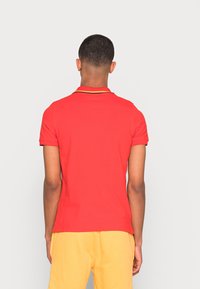 Czerwony polo z dopasowanym krojem, z żółtymi wykończeniami kołnierzyka i krótkimi rękawami. Tło jest jednolicie białe.