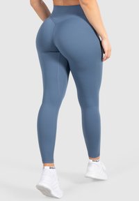 Leggings a vita alta in tessuto blu, con una texture liscia e un design aderente. Presentano un logo discreto e scarpe da ginnastica bianche.