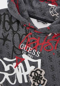 Mikina s kapucí ve tmavě šedém materiálu s grafiky ve stylu graffiti v červené a bílé barvě, včetně loga "GUESS EST. 1981" a vzorovaných akcentů.