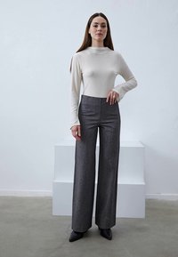 Helles beiges Langarmoberteil kombiniert mit hoch taillierten, weit geschnittenen grauen Hosen. Glattes Material mit einer figurbetonten Silhouette. Schwarze spitze Schuhe.