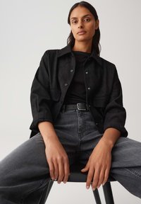 Giacca nera in suede con bottoni, indossata sopra un top nero a coste, abbinata a jeans grigi a vita alta. Modella seduta su una sedia nera.