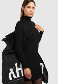 Mustast soonilisest ribikoega kleit kõrge kraega, mille peal on avatud tõmblukuga must sulejakk, mille siseküljel on valge logo.