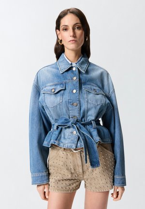 Donna con giacca di jeans blu con bottoni frontali e cintura in vita, abbinata a pantaloncini beige decorati con borchie metalliche.