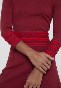 Femme en robe à manches longues bordeaux avec des poignets et une ceinture rayés rouges, les bras croisés à la taille.