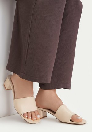 Piedi con sandali beige aperti con tacco largo abbinati a pantaloni corti marroni testurizzati su sfondo bianco.