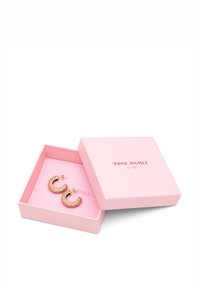 Øreringe i guldton med lyserøde ædelstens detaljer, præsenteret i en lyserød æske med mærket "TONE DAMLI" trykt på.