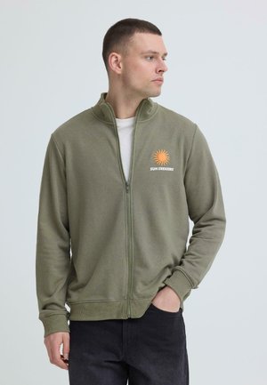 Mężczyzna w oliwkowozielonej kurtce na zamek z pomarańczowym logo słońca i napisem „SUN SEEKERS”, patrzący w lewo, z jedną ręką w kieszeni.