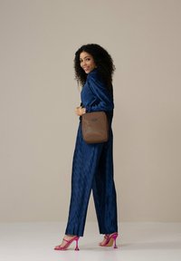 Braune, strukturierte Umhängetasche in quadratischer Form, die über einem blauen, gefalteten Outfit getragen wird und mit rosa Absatzsandalen kombiniert ist.