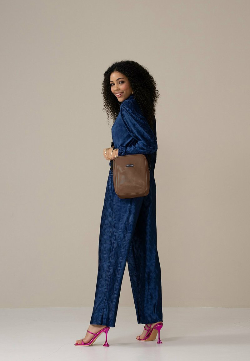 Braune, strukturierte Umhängetasche in quadratischer Form, die über einem blauen, gefalteten Outfit getragen wird und mit rosa Absatzsandalen kombiniert ist.